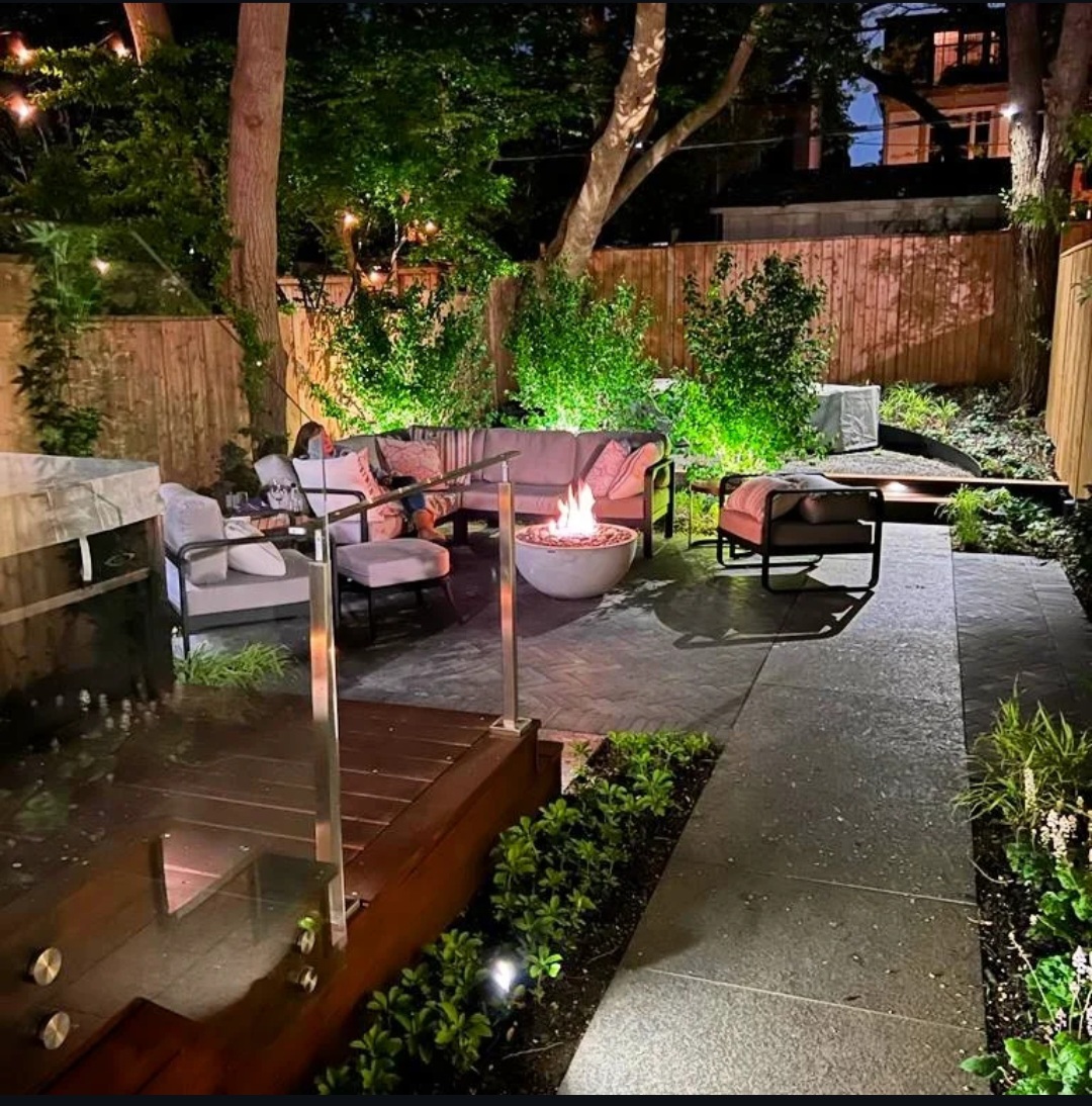 The finished garden at night with the fire bowl lit and the planting uplit
