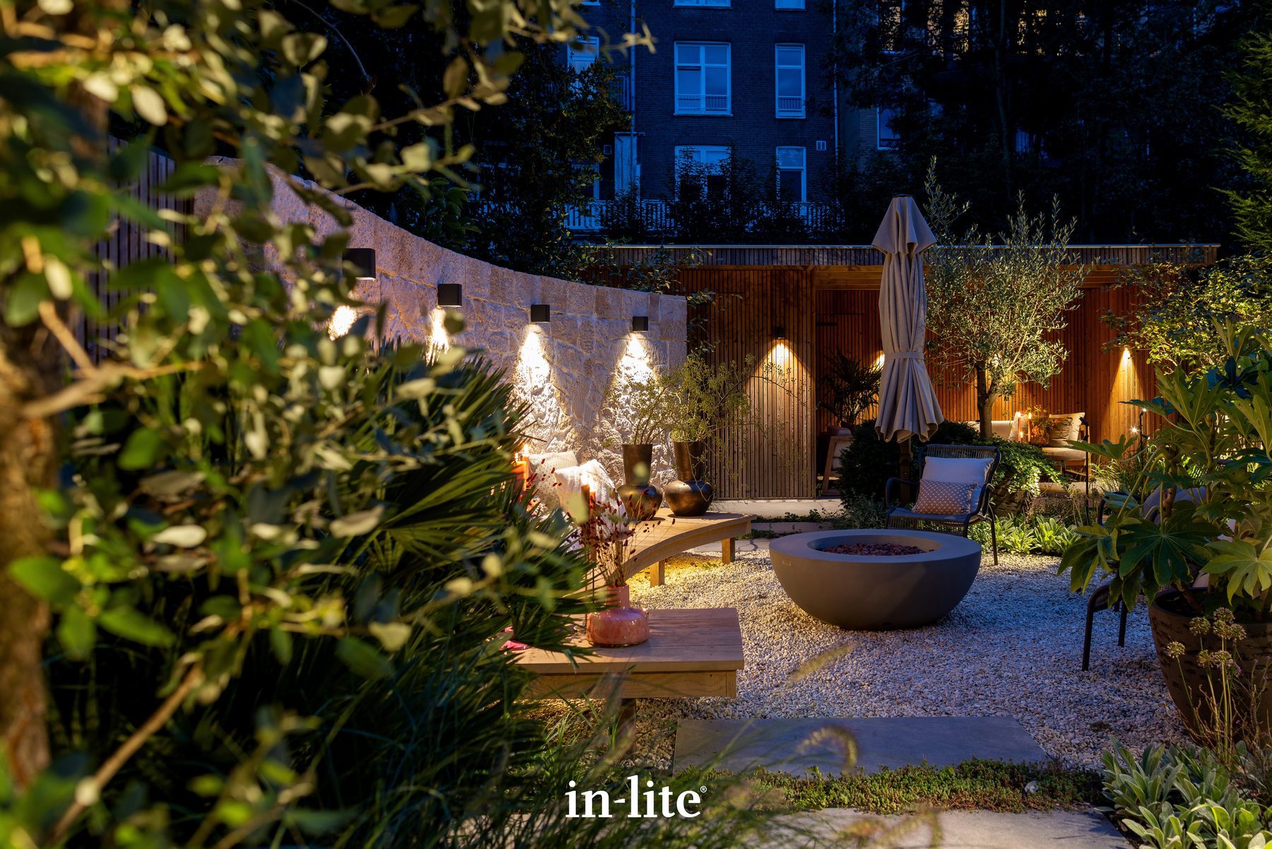 Planted courtyard after dark, wall lights washing a curved stone wall with an olive tree and fire bowl beyond