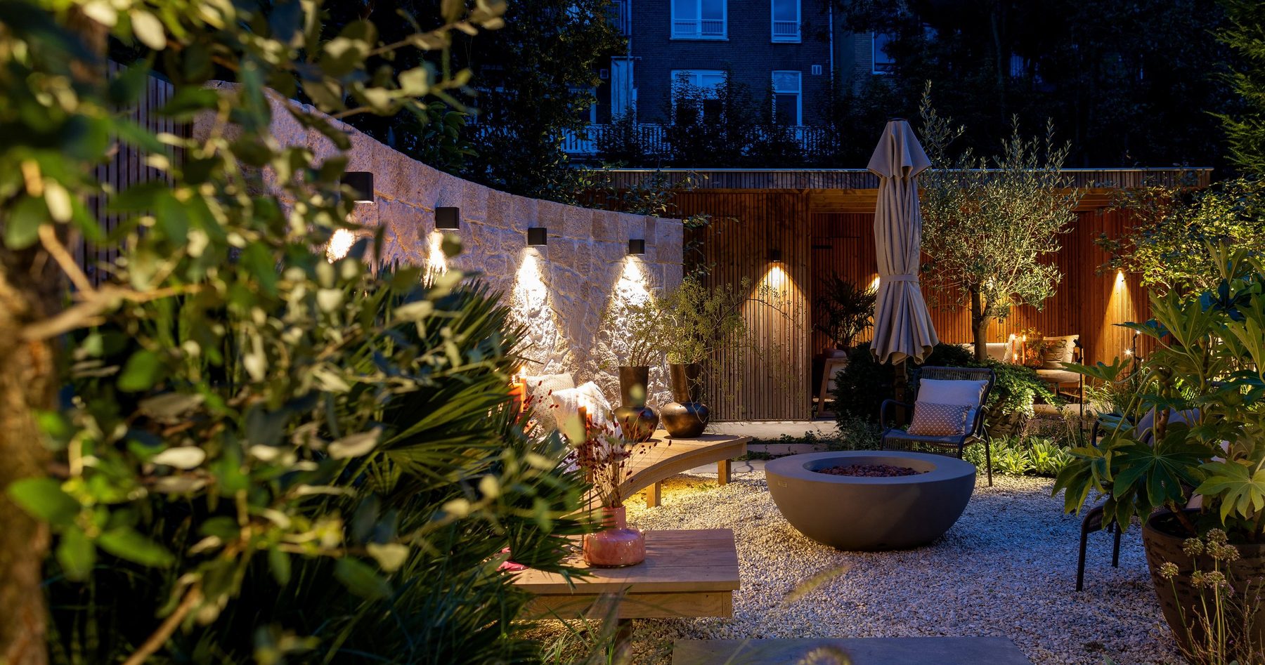 Courtyard garden after dark, wall lights grazing stone and a lit fire bowl